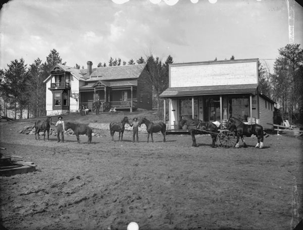View down unpaved road towards a frame house and storefront. In the background in the yard of the house is a man standing and a woman sitting with a child. In the foreground two men are displaying four horses in the road. On the right is a man driving a horse pulling a buggy, with another horse tied behind.
