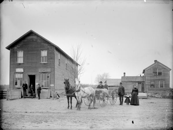 Exterior view of post office/store and a frame house in the town of Shamrock, Two men and a boy are standing in front of the store, a man and woman are standing and holding the hands of a girl in front of the house, and in the center are a man and woman are sitting in a buggy pulled by two horses.