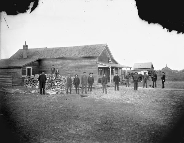 Group of men and boys posed standing in front of a wooden building, probably a tavern. The photographer's wagon is in the background.