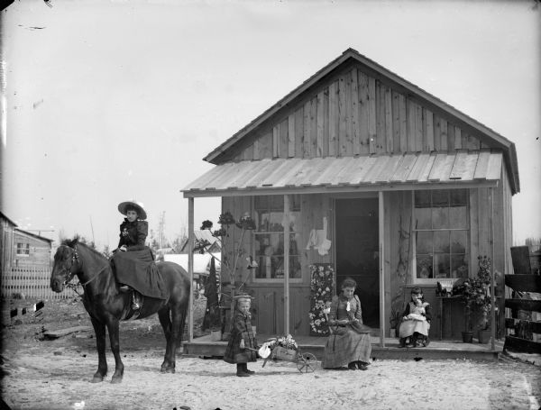View of a woman posing sitting on the porch of a wooden building and sewing a hat, probably a milliner in front of her shop. A small girl is sitting in a rocking chair on the porch holding a doll, and another small child is pushing a wheelbarrow full of flowers. Another woman on the far left is sitting sidesaddle on a horse. There is a tree branch on the porch holding a display of hats.