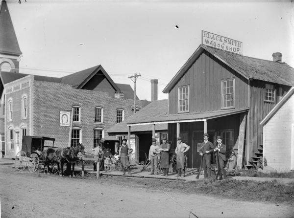 View across street towards six men posing, with four of the men wearing aprons and work clothes, and two men in suits holding guns. Beside them on the left is a woman and girl standing next to a baby in a baby carriage. The group is standing in front of a blacksmith and wagon shop. The photographer's wagon with a team of horses is parked along the sidewalk on the far left. On the extreme left in the background is a brick building, and a steeple behind rising above a second building.