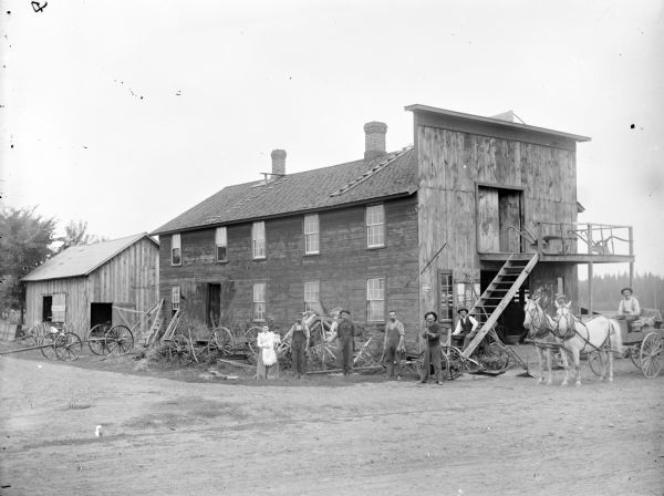 Group portrait of five men, and a woman holding a baby, standing in front of a large, two-story wooden building, possibly a farm implement or feed store. Another man is sitting in a wagon pulled by two horses. A sign on a shed on the left reads: "Universal Plows," and two plows are displayed on a landing above the horse-drawn wagon.