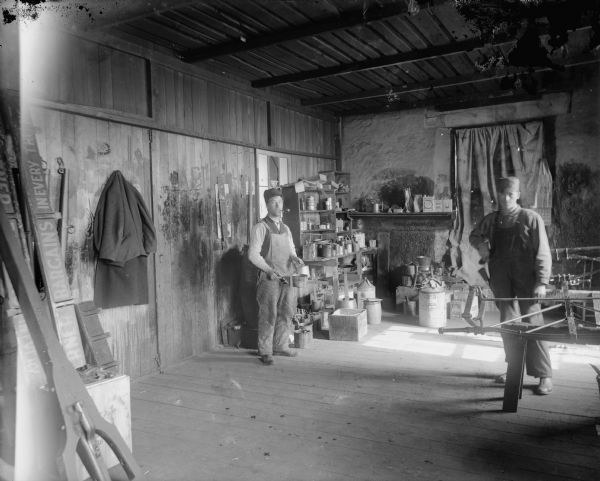 Painters posed standing in a workshop near a wagon frame. The man on the left has a pipe in his mouth.