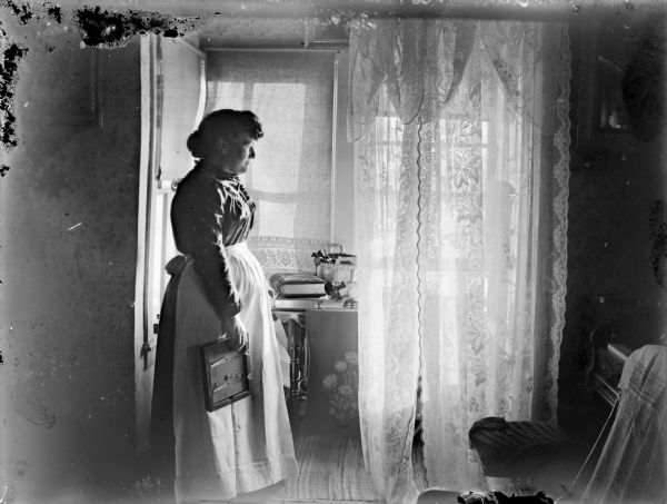 Woman posed standing by a window and table in a home. She is holding an object in her right hand, possibly a frame for photographs. On the far right is a piano partially covered by a cloth.