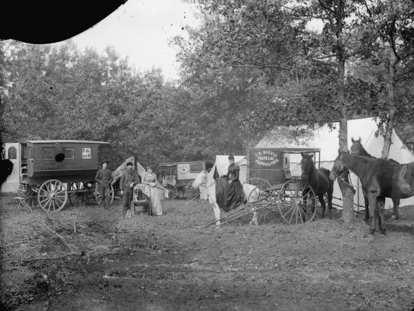 Three photographer's wagons, two owned by C.R. Monroe and the other by N.L. Ellis, in front of tents in the countryside. Two men and a woman are posing standing by a chair in which a dog is sitting. On the right is a woman mounted sidesaddle on a horse. The man and the woman by the chair may be C.R. Monroe and his wife, and the woman on the horse may be their daughter. The man on the far left may be N.L. Ellis. Three other horses are tethered on the far right in front of a tent.