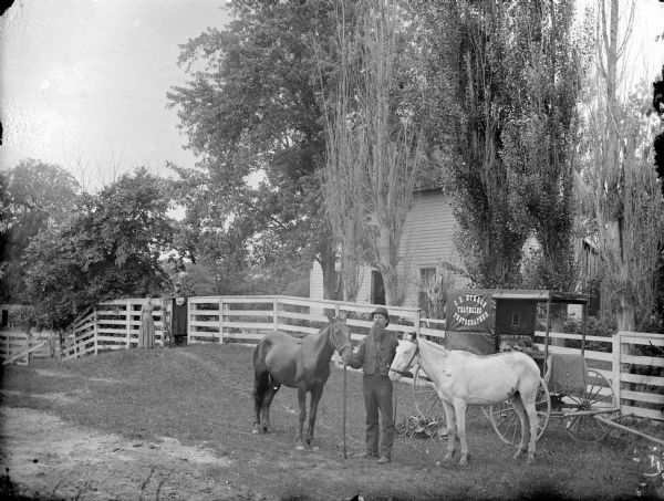 Man displaying two horses in front of a photographer's wagon that says "C.R. Monroe, Traveling Photographer." Two women, one holding a photograph, are posed standing by the gate of a wooden fence and a frame house in the background. The group may be C.R. Monroe, and his wife and daughter.