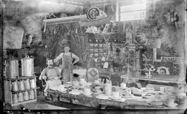 Man posed seated and another man standing in a large room, perhaps a shop of some kind, at a table set for a large meal. Both men are wearing aprons. On their left is a shelf with two large beverage dispensers, with pitchers stored below. Above them and in the background are a curtain, with banners, flags and other decorations. On the right is a large assortment of photographs and posters behind a glass display case with a cake stand full of apples.