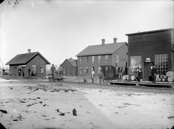 Men at a railroad station awaiting the arrival of a train in Fremont (later Pray). There is snow on the ground. Two men are standing on a hand-powered rail cart on the railroad tracks. Some of the men waiting for the train are carrying tools. A sign for the Western Union Telegraph Office is on the building on the left. The building on the right has a Post Office sign above the door.