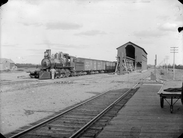 Two men posed by the locomotive of a freight train on railroad tracks, probably by the coal shed at Millston. The two-wheeled express wagon on the platform was for hauling items in and around the freight house.