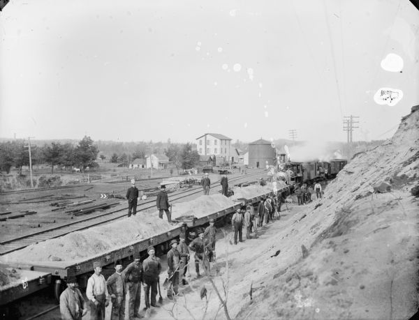 View from steep bank of a large group of men posed standing with shovels by a railroad train with flat cars covered with sand or dirt. Man of the men are holding shovels, and standing at the foot of a steep embankment. In the background are piles of lumber, as well as houses and a large three-story building.