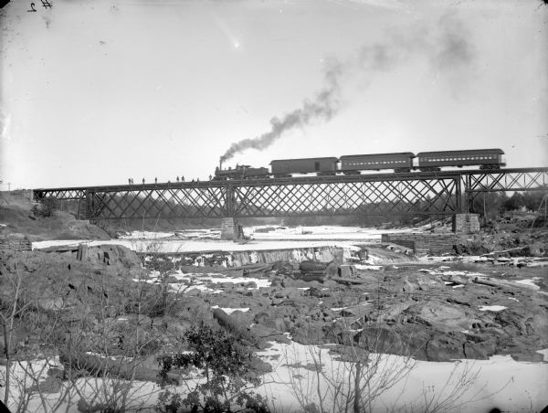 View from shoreline of a passenger train on a railroad bridge over a river. Men are standing on the bridge in front of the locomotive. There is snow on the ground and ice on the river.