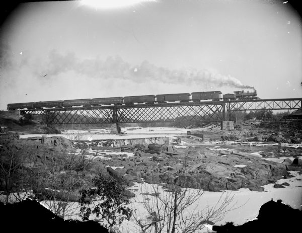 View from shoreline of a passenger train on a railroad bridge over a river. There is snow on the ground and ice on the river.