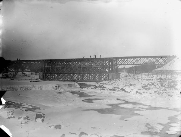 View from shoreline over river of men posed standing on a railroad bridge under construction during the winter.