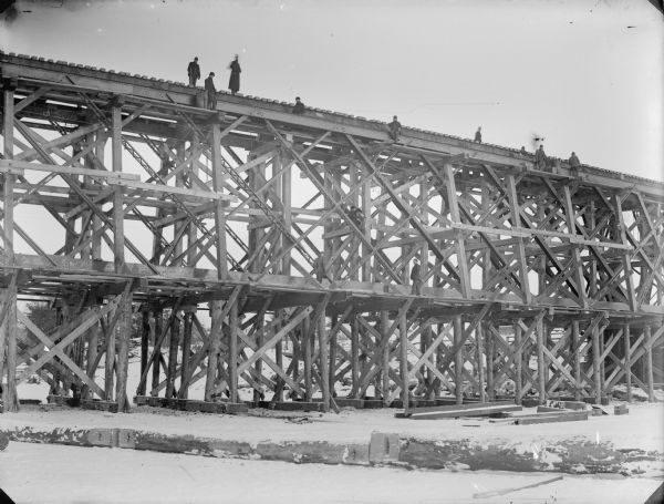 View from frozen river of men posed high above on a railroad bridge under construction during the winter. There is ice on the river and snow on the ground.