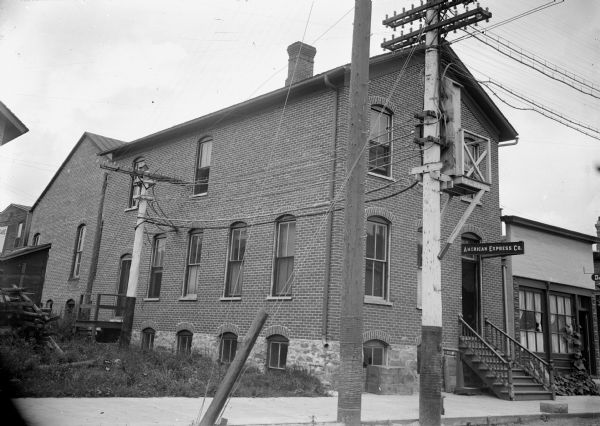 Exterior view from street of brick two-story building partially owned by Charles J. Van Schaick which housed the American Express Company and Exchange Office of the Central Wisconsin Telephone Company. Van Schaick had his photography studio upstairs.