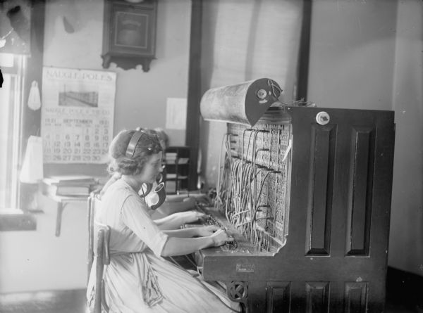 Two woman operators posed seated at the Kellogg 350 drop switchboard of the telephone exchange. Calendar on the wall shows a date of September 1921.