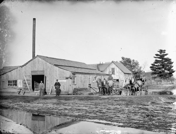 Two men posed standing outside a wooden building with a tall smokestack, probably a power plant for a sawmill. Two men are posed sitting in two wagons, each pulled by teams of two horses, parked by the building on the muddy ground.