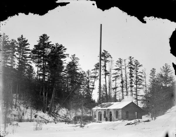 Three men posed standing on the porch of a small brick building with a tall smokestack, probably the City Water Pumping Station at Town Creek, surrounded by snow-covered ground.