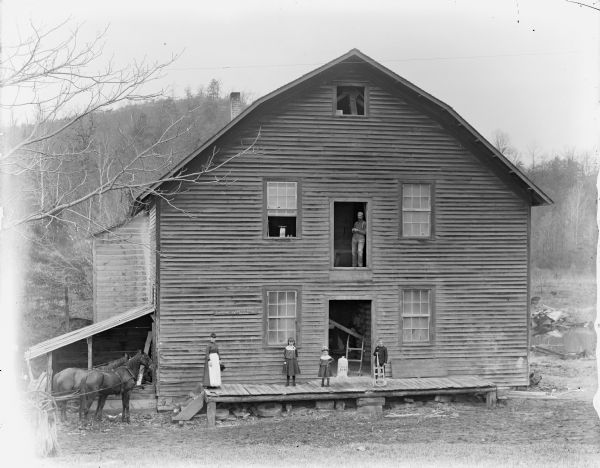 view of a woman, two girls, and a boy posed on the porch of a two-story wooden building, probably the John Edmunds' Charter Oak grist mill built on Squaw Creek in 1866. Above them in an open doorway a man is posed standing. The mill was owned and operated by George Washington Cooper, the father of George Cooper, an engineer who built the original Dnieprostroi Dam in the Soviet Union. The boy may be George Cooper. A small sign on the building reads: Spaulding. Takes the Lead. Wagons."