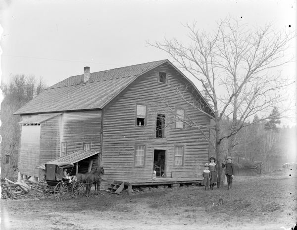 Two girls and a boy posed by a tree in front of a two-story wooden building, probably the John Edmunds' Charter Oak grist mill built on Squaw Creek in 1866. On the left a woman is posed sitting in a buggy pulled by two horses. A man os visible through the doorway. The mill was owned and operated by George Washington Cooper, the father of Hugh Cooper, an engineer who built the original Dneiperstroy Dam in the Soviet Union. The boy may be Hugh Cooper. A small sign on the building reads: Spaulding. Takes the Lead. Wagons."