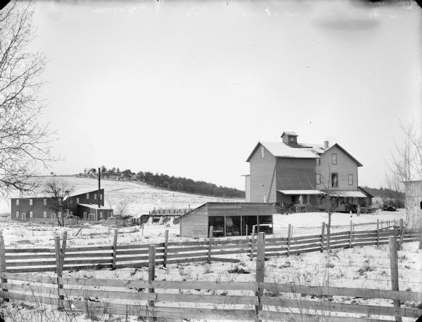 View across fields and fences of three wooden buildings in the country, probably a mill. A man stands in the wagon drawn by a team of horses in front of one of the three-story building. Another man stands in the open doorway on the second floor. Below him another man sits in a wagon, and in the snowy yard, a young girl and a young man stand. In the far background is a hill.