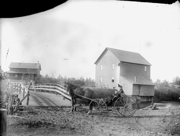 Woman and girl posed sitting under a fur in a carriage pulled by a single horse by a wooden bridge. Behind them is a large wooden building, possibly a mill on Robinson Creek owned by Moses Paquette. In the background beyond the bridge is a house with a front porch.