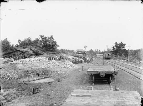 Group of men at the construction of a railroad branch line among piles of stones. Railroad ties placed on the ground are visible on the platform in the background. Photograph taken from atop a flat car.