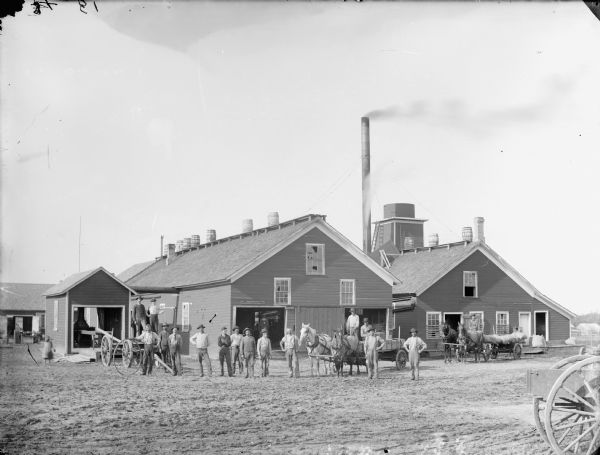 Group of men and a young girl posed standing and in wagons outside two large wooden buildings, possibly a sawmill. There is a row of barrels along the top of the roof of the center building.