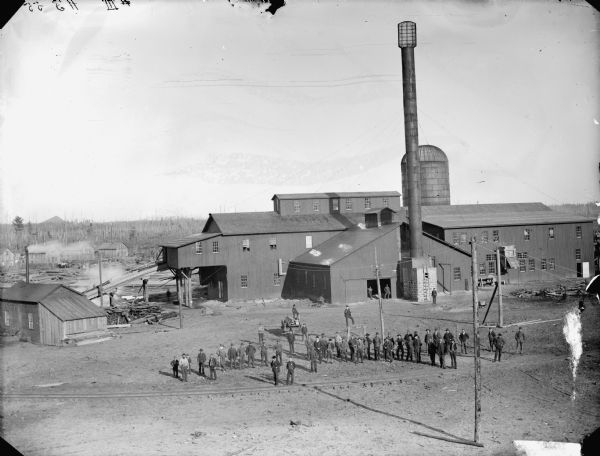 Elevated view of a large group of men posing standing in front of a sawmill, probably the Goodyear Mill, three miles south of Saddle, or a mill located in McKenna.