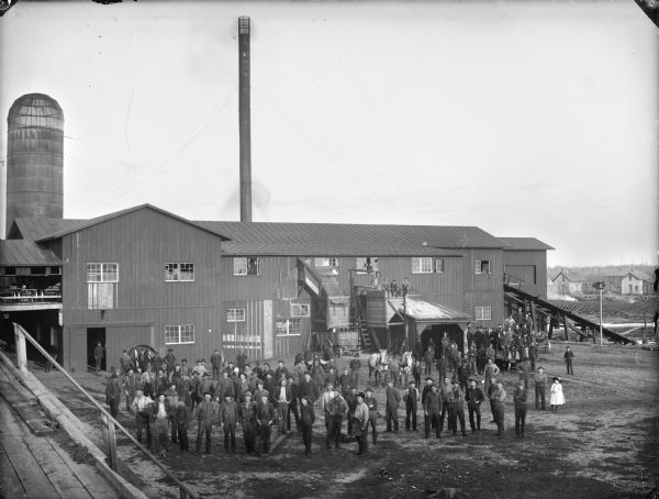 Elevated view of a large group of men, children and horses posed standing in front of a sawmill, probably a mill located in McKenna.