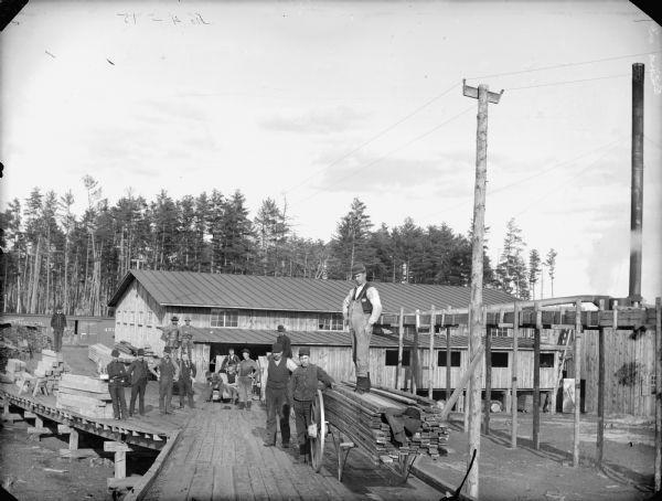 Large group of men posed standing near and on a wagon stacked with timber which is on top of a platform in front of a sawmill. Railroad cars are behind the long building in the background. Probably a newly-constructed sawmill in McKenna.