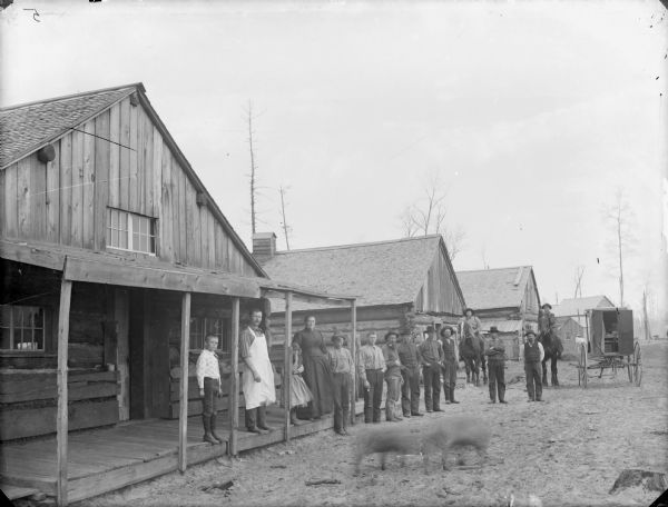 View from road of several men, a woman, girl and young boys posed in front of buildings in a lumber town, probably McKenna. There are two blurred images in the foreground, which are probably dogs or pigs.