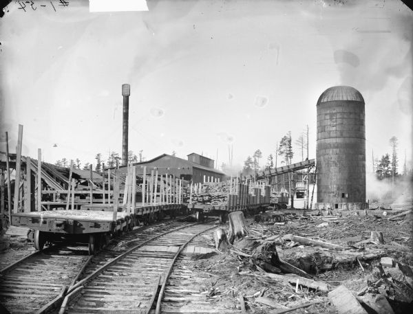 View down railroad tracks entering a lumberyard, with a silo on the right, and chimney on the left. Possibly the Goodyear Mill, three miles south of Saddle. Or possibly a mill located in McKenna.