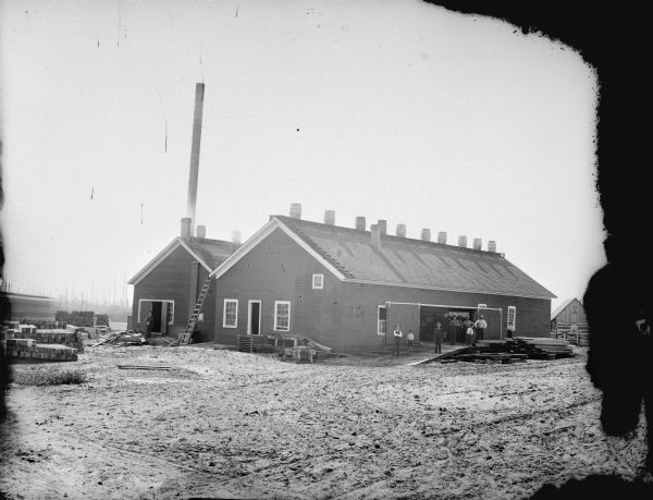 View across yard of men and boys posing in the doorway of a sawmill. There is a row of barrels across the top of the roof.