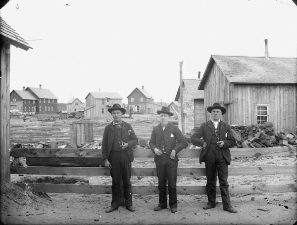 Three men posed standing against a fence holding pipes in a yard. In the background is a river clogged with floating logs floating. Several wooden buildings are on the far shoreline.