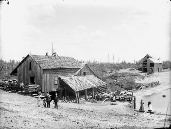 View down hill of three women, three men, and a boy and girl posed standing in different locations around a sawmill by a shoreline, possibly the Lath Mill. The girl is holding two cats.
