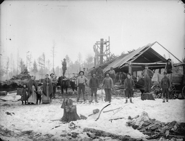 Large group of men, women, and children posed standing on the snow-covered ground in front of a sawmill either under construction, or an open saw mill.