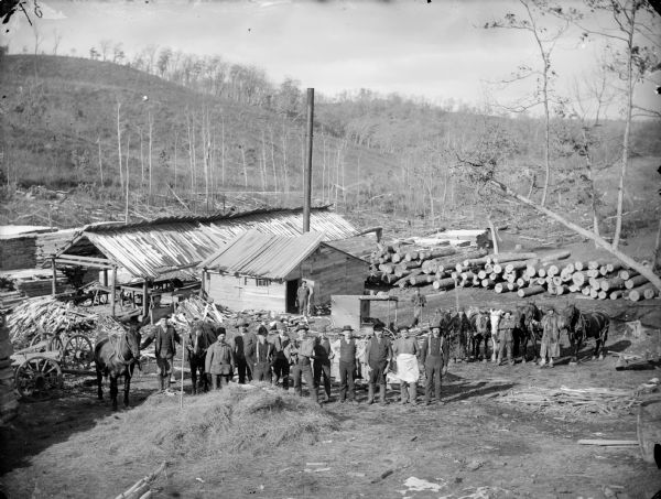 View down hill of a large group of men, some displaying horses, in front of a sawmill and its power plant. One of the men is holding a dog, and two of the men wear long fur coats.