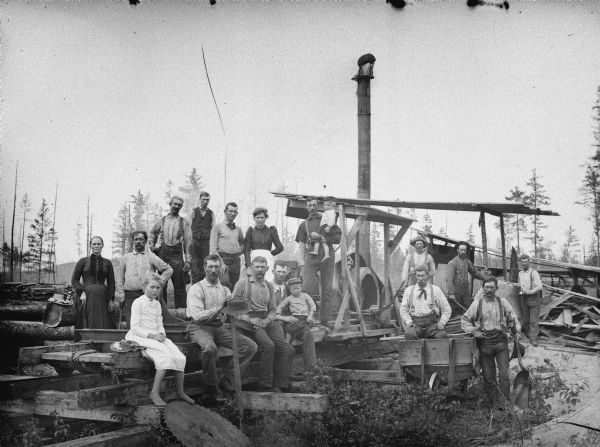 Large group of men, women, and children posed standing and sitting in front of an open sawmill.