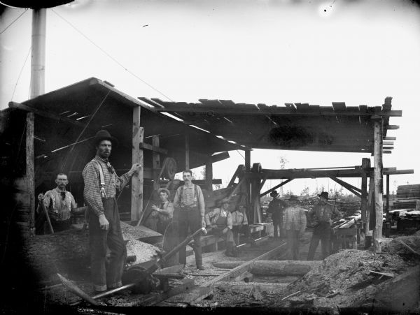 Men posed standing and sitting near the saw blade of an open sawmill.