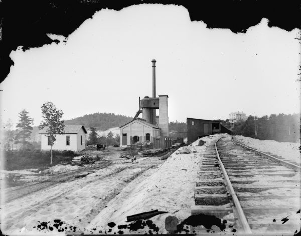 York Iron Works with a view of the blast furnace. On the hill is the boardinghouse and the dance hall. A man is working near the railroad cars, and other people are standing in the open doorway of the blast furnace building.