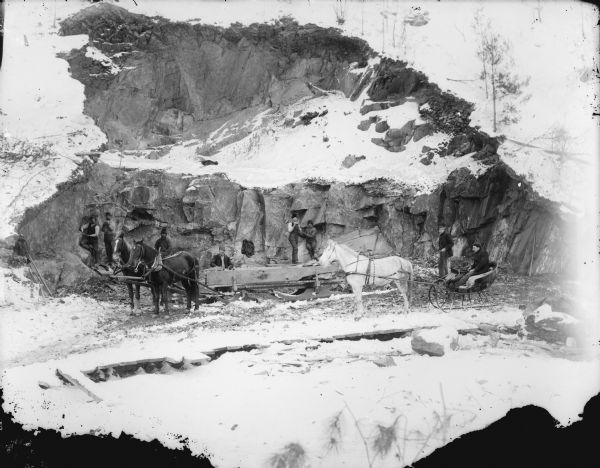 View down hill into quarry with men posed working the rock face behind a bobsled pulled by a team of two horses. A man is sitting in a cutter sleigh pulled by a single horse.