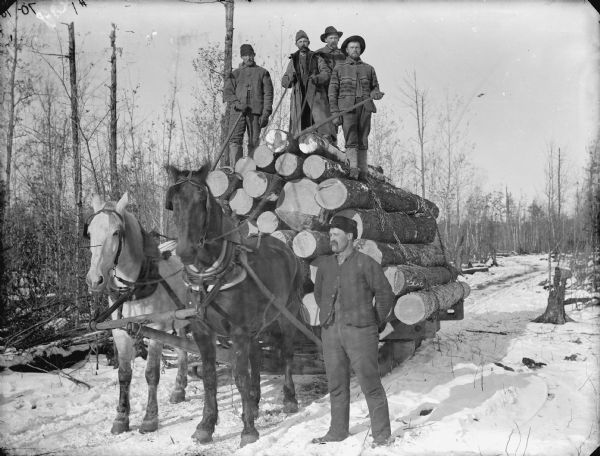 Four men posed with lumber tools atop a load of logs on a bobsled pulled by a team of two horses. A man is standing near the team, probably James Abram Bailey.