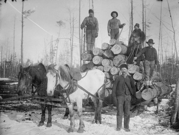 Four men posed with lumber tools atop a load of numbered logs on a bobsled pulled by a team of two horses. A man is standing near the team, probably James Abram Bailey.	