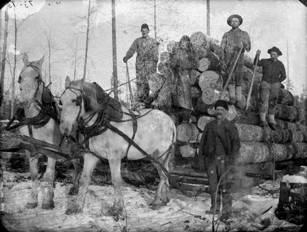 Four men posed with lumber tools atop a load of numbered logs on a bobsled pulled by a team of two horses. A man is standing near the team, probably James Abram Bailey.	