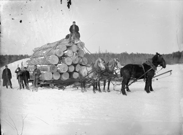 Man posed sitting on a bobsled loaded with logs and pulled by a team of four horses. Another four men are holding logging tools posed next to the bobsled.