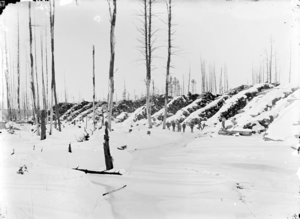 Group of men and children posed in front of large piles of snow-covered logs, ready to be dumped from landings into the river after the spring thaw.	