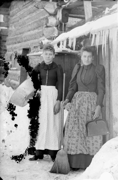 Two women stand outside the doorway of a log house in winter. They are posed with a washtub, broom and dust pan.