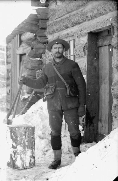 A man posing with an ax and chopping block is standing outside the door of a log cabin during the winter. The man has been identified as James B.F. Server, who was married to Dora Van Buskirk. She was the eldest child of John Van Buskirk and Elizabeth.
  