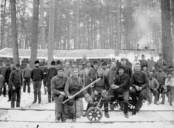 Loggers posing on narrow-gauge railroad tracks with a railroad velocipede and cant hook near Saddle Mound, a large hill that dominates the local area in Jackson County. There is a long, low building behind them.